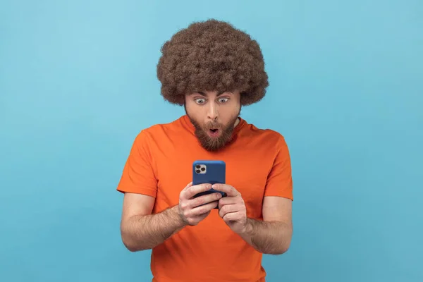 携帯電話を使用してソーシャルネットワーク上のオレンジ色のTシャツの読書の投稿を着てアフロの髪型と驚きの男の肖像画 驚いて見てチャット 青い背景に隔離された屋内スタジオショット — ストック写真