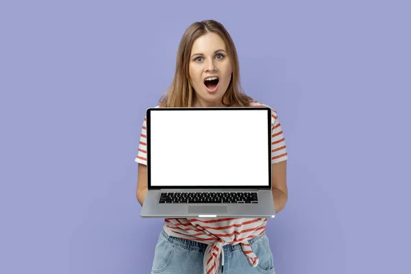 広告のための空のスペースと白い空のディスプレイとノートパソコンを保持縞Tシャツを着てショックを受けた金髪の女性の肖像画 紫色の背景に隔離された屋内スタジオショット — ストック写真