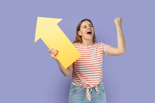 縞模様のTシャツを着て非常に幸せな女性の肖像は大きな黄色の矢印を脇に指していると拳を握りしめ 勝利を祝う 増加します 紫色の背景に隔離された屋内スタジオショット — ストック写真