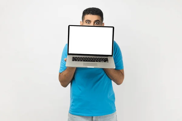 害羞的年轻人穿着蓝色T恤站在笔记本电脑后面 用空的显示屏遮住半张脸 看着相机的画像 在灰色背景下被隔离的室内拍摄 — 图库照片