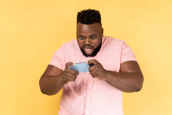 stock image Portrait of amazed man wearing pink shirt standing, using smartphone and playing mobile game with excited face and big eyes. Indoor studio shot isolated on yellow background.