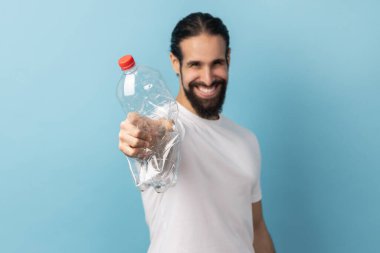 Beyaz tişört giyen, plastik şişe tutan, plastik geri dönüşüm ve çevre kirliliğinden endişelenen sakallı bir adamın portresi. Mavi arka planda kapalı stüdyo çekimi.