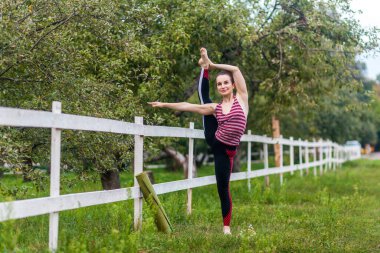 Bacağını esneten, yoga yapan, dışarıda antrenman yapan, havada bacak kaldıran, bir ayağını ikiye ayıran, gülümseyerek uzaklaşan esnek ve güçlü genç bir kızın tam boy portresi..