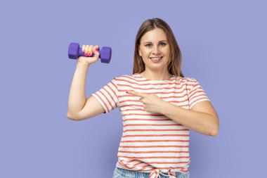 Elinde dambıl tutan çizgili tişört giyen, bisepsini gösteren ve gülümseyen zevkli sarışın kadının portresi. Mor arka planda izole edilmiş kapalı stüdyo çekimi.