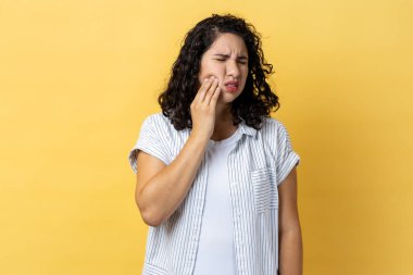 Sağlıksız bir kadın portresi. Koyu dalgalı saçlı, yanağına dokunan ve acı içinde kıvranan korkunç diş ağrısı çeken periodontal bir hastalık. Sarı arka planda kapalı stüdyo çekimleri.