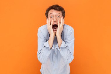 Kameraya ağzı açık bakan korkmuş bir adamın portresi, elleri yanaklarda, korkmuş, açık mavi gömlekli. Turuncu arka planda kapalı stüdyo çekimleri.