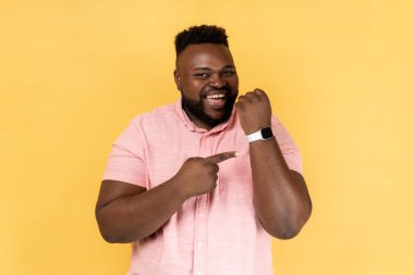 Zaman doldu. Pembe gömlek giyen ve kameraya bakıp akıllı saatini gösteren mutlu, sakallı bir adamın portresi. Sarı arka planda kapalı stüdyo çekimleri.