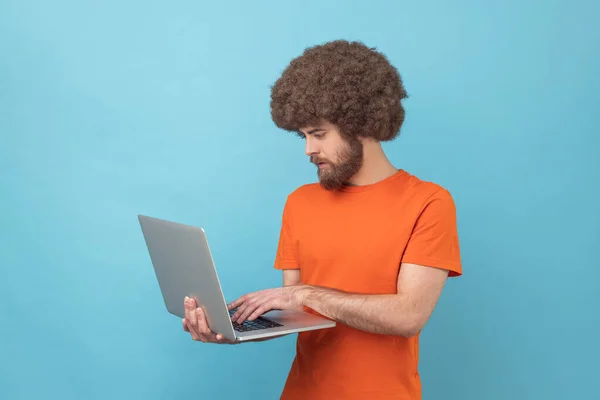 手にノートパソコンを持ち コンピューターで作業し 真剣な表情でディスプレイを見ているオレンジ色のTシャツを着たアフロヘアの男のサイドビュー 青い背景に隔離された屋内スタジオショット — ストック写真