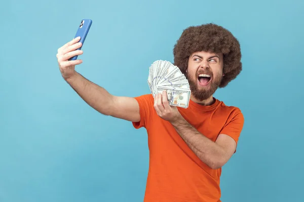 ビデオ通話やライブストリームを持つオレンジ色のTシャツを着てアフロの髪型を持つ興奮した男の肖像画は 加入者にドル紙幣を示しています 青い背景に隔離された屋内スタジオショット — ストック写真