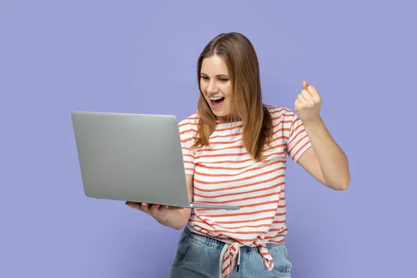 非常に幸せな美しい金髪の女性の肖像画ストライプのTシャツノートパソコンで作業を着て 画面を見て 拳を握りしめ はいジェスチャーを示しています 紫色の背景に隔離された屋内スタジオショット — ストック写真
