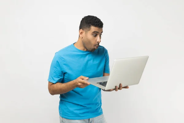 穿着蓝色T恤衫站在那里 手里拿着笔记本电脑 在网上工作 睁大了大了眼睛 惊讶地看着屏幕 在灰色背景下被隔离的室内拍摄 — 图库照片