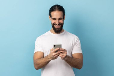 Sakallı, beyaz tişörtlü, akıllı telefon kullanan, cihaz ekranına bakıp yazı yazan yakışıklı, pozitif adam portresi. Mavi arka planda kapalı stüdyo çekimi.
