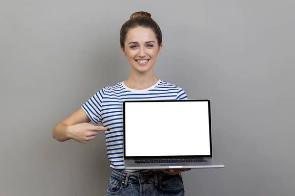 白い空のディスプレイをラップトップで指を指して縞Tシャツを着ている女性の肖像画は 空の画面を示し インターネット広告で自慢します 灰色の背景に隔離された屋内スタジオショット — ストック写真