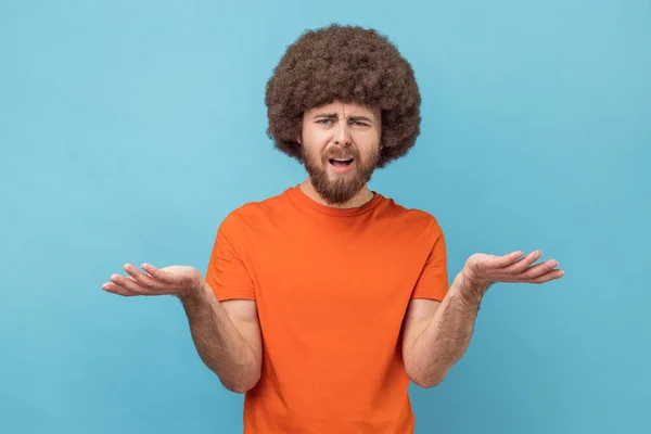 Portrait of stressed annoyed man with Afro hairstyle wearing orange T-shirt standing raised arms and asking, expressing negative emotions. Indoor studio shot isolated on blue background.