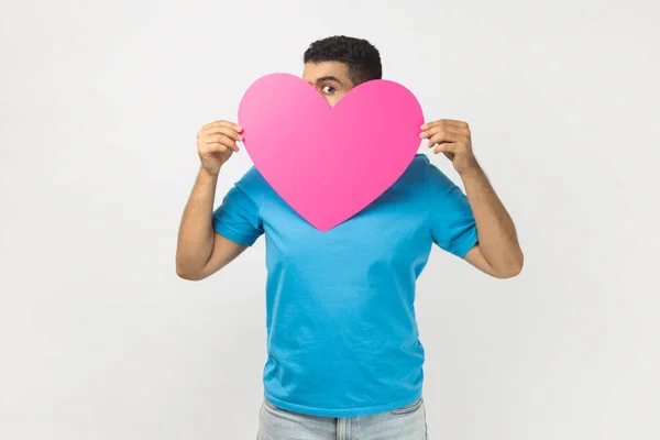 害羞的匿名男子穿着蓝色T恤站在大大的粉色心脏里窥视 羞于在情人节表达自己的感受的画像 在灰色背景下被隔离的室内拍摄 — 图库照片
