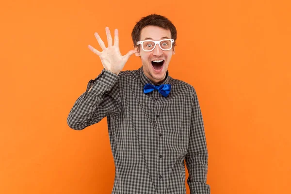 Portrait of funny excited man nerd waving his hand, showing hello or bye gesture, wearing shirt with blue bow tie and white glasses. Indoor studio shot isolated on orange background.