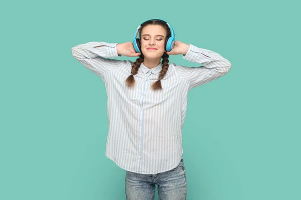 ヘッドフォン 閉じた目と笑顔で好きな音楽を聞いて立って縞模様のシャツを身に着けている編組とティーンエイジャーの女の子の肖像画 緑の背景に隔離された屋内スタジオショット — ストック写真