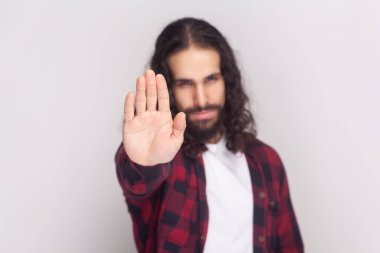 Kırmızı tişörtlü, uzun kıvırcık saçlı ciddi sakallı adam avuç içi ile dur işareti yapıyor, yasaklama ve yasaklama gösteriyor, bir şeyi reddediyor. Gri arka planda izole edilmiş kapalı stüdyo görüntüleri.