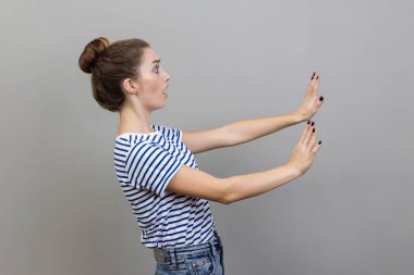 Çizgili tişört giyen korkmuş genç bir kadının yan görünüş portresi. Avuç içleriyle hareket etmeyi kes ve korkmuş gözlerle şaşırmış görün. Gri arka planda izole edilmiş kapalı stüdyo görüntüleri.