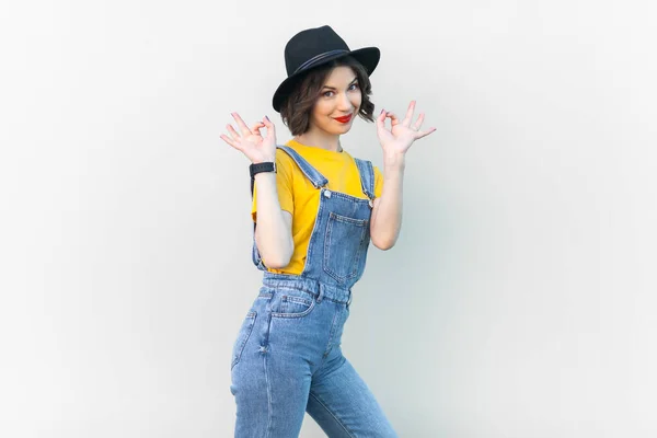 青いデニム全体で楽観的な幸せなヒップスター女性の肖像画 黄色のTシャツと黒の帽子は 彼女の腕を上げ 大丈夫なサインを示しました 灰色の背景に隔離された屋内スタジオショット — ストック写真