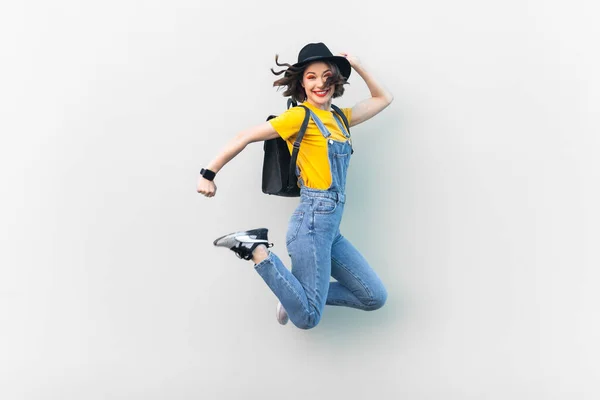 ブルーデニム全体で魅力的な喜びのヒップスター女性の肖像画 黄色のTシャツと黒の帽子 ジャンプ 気分が良い 幸福を表現している 灰色の背景に隔離された屋内スタジオショット — ストック写真