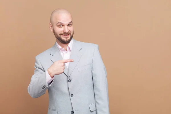 笑容满面的秃头男子站在广告区 复印促销空间 身穿灰色夹克 在棕色背景下被隔离的室内拍摄 — 图库照片