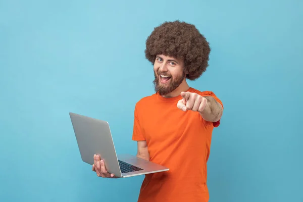 手にポータブルコンピュータで立ってオレンジ色のTシャツを着てアフロの髪型を持つ幸せな男の肖像画は カメラを見て 指しています 青い背景に隔離された屋内スタジオショット — ストック写真