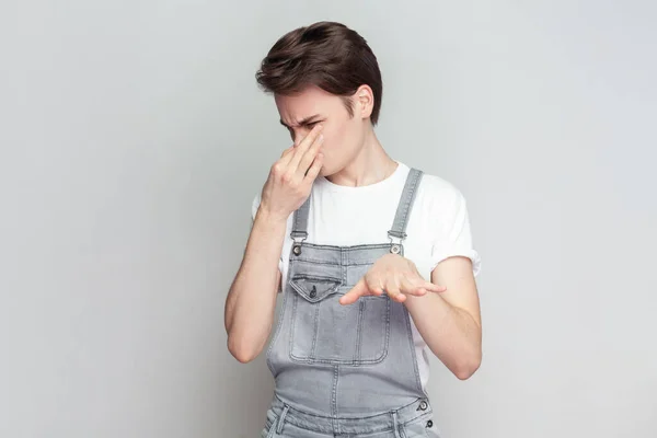 Young brunette man covers nose with hand, smells something awful, pinches nose, frowns in displeasure, sees pile of garbage, wearing denim overalls. Indoor studio shot isolated on gray background.