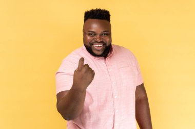 Yanıma gel. Pembe gömlekli bir adamın portresi, işaret edici jestler yapıyor, randevu için davet ediyor ve çekici görünüyor, kameraya flört ediyor. Sarı arka planda kapalı stüdyo çekimleri.