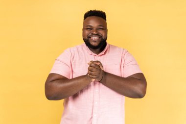 Mutlu, mutlu, mutlu, sakallı, pembe tişörtlü bir adamın portresi ayakta duruyor, el ele tutuşuyor, dişlek bir gülümsemeyle kameraya bakıyor. Sarı arka planda kapalı stüdyo çekimleri.