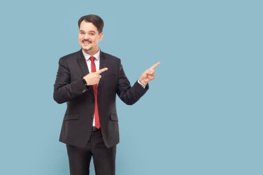Gülümseyen, bıyıklı, reklam veya tanıtım metni için boş yeri işaret eden, kırmızı kravatlı, siyah takım elbiseli olumlu bir adam. Açık mavi arkaplanda izole edilmiş kapalı stüdyo çekimi.