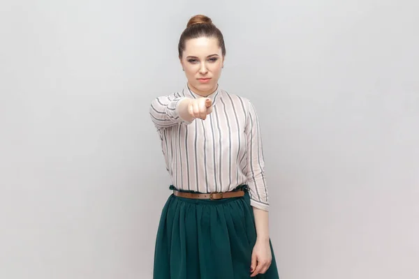 Portrait of serious strict bossy woman wearing striped shirt and green skirt standing pointing to camera, choosing you, scolding. Indoor studio shot isolated on gray background.