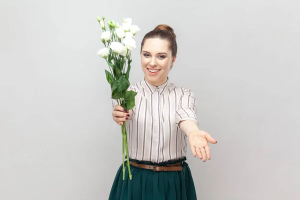 白花の花束を保持縞模様のシャツと緑のスカートを身に着けている陽気なかなり魅力的な女性の肖像画は 手を広げ あなたのためにそれを与えます 灰色の背景に隔離された屋内スタジオショット — ストック写真