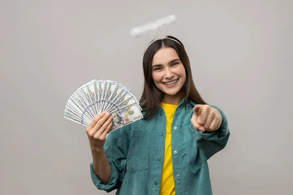 頭の上にハローを持つ幸せな天使の若い女性はドル紙幣を保持し カメラを指して 宝くじの勝者を選択し 大きなお金を稼ぐことを奨励する 灰色の背景に隔離された屋内スタジオショット — ストック写真