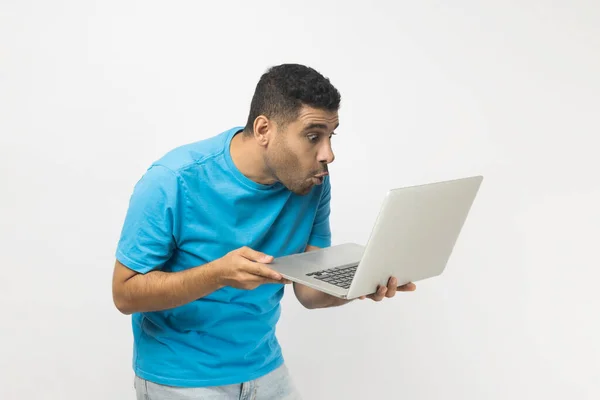 穿着蓝色T恤的不修边幅的男人吃惊地站在电脑屏幕前 用大大的眼睛望着监视器 看到了令人惊讶的东西 在灰色背景下被隔离的室内拍摄 — 图库照片