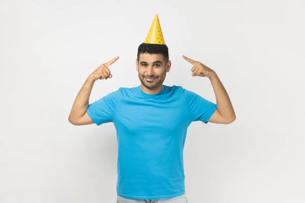 青いTシャツを着て黄色のパーティーコーンを指して立っているお祝いの肯定的な楽観的なUnshaven男の肖像画は 彼の誕生日を祝います 灰色の背景に隔離された屋内スタジオショット — ストック写真