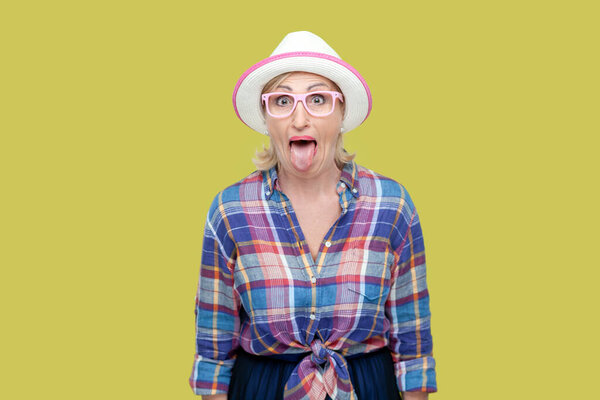 Портрет взрослой женщины в клетчатой рубашке, шляпе и очках, торчащей из языка, делающей смешную гримасу. Крытая студия снята на жёлтом фоне.