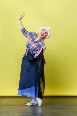 Şapka ve gözlük takıp kameraya mutluluk ve dansla bakan mutlu, yaşlı bir kadının tam boy portresi. Sarı arka planda kapalı stüdyo çekimleri