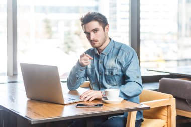 Dizüstü bilgisayarda çalışan ve projeyi bitirmen gerektiğini söyleyerek kamerayı işaret eden, kot pantolonlu, serbest çalışan bir gencin portresi. Büyük pencerenin yanında, kafenin arka planında..