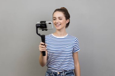 Çizgili tişört giyen gülümseyen kadın blogcunun portresi Steadicam ve cep telefonunu kullanarak takipçilerle konuşuyor. Gri arka planda izole edilmiş kapalı stüdyo görüntüleri.