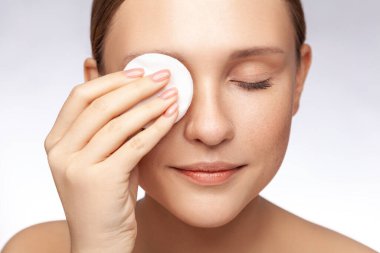 Kapalı gözlü, çıplak duran, yüzüne pamuk pedleri gösteren, duştan önce makyajını silen güzel bir kadının yakın plan portresi. Kapalı alanda çekilen çekim gri arka planda izole edildi.