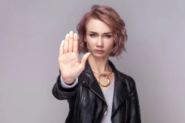 Portrait of strict bossy beautiful woman with short hairstyle standing looking at camera, showing stop sign, wearing black leather jacket. Indoor studio shot isolated on grey background.