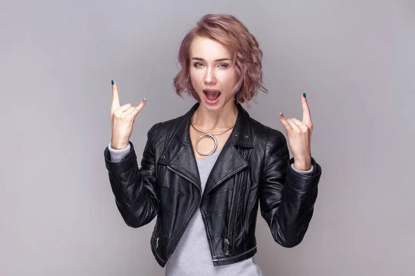 Crazy funny woman with short hairstyle standing with rock and roll gesture, heavy metal sign, having fun on festival, wearing black leather jacket. Indoor studio shot isolated on grey background.