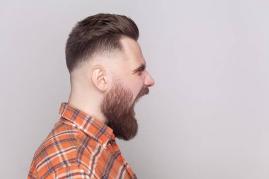 Ciddi derecede sert sakallı bir adamın portresi. Önüne bakıyor, saldırganlıkla bağırıyor, turuncu kareli gömlek giyiyor. Gri arka planda izole edilmiş kapalı stüdyo görüntüleri.