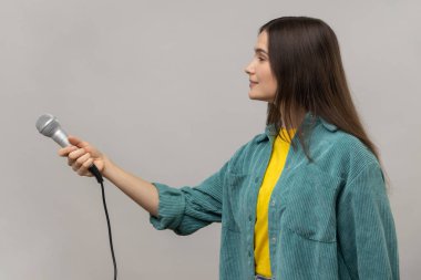 Koyu saçlı, mikrofon sunan gazeteci ya da muhabirin sıradan bir ceket giyen tatmin olmuş bir kadının yan görüntüsü. Gri arka planda izole edilmiş kapalı stüdyo görüntüleri.