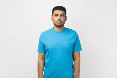 Tıraşsız, ciddi derecede yakışıklı bir adamın portresi. Mavi tişört giymiş, kameraya bakıyor ve konsantre bir ifadeye sahip. Gri arka planda izole edilmiş kapalı stüdyo görüntüleri.