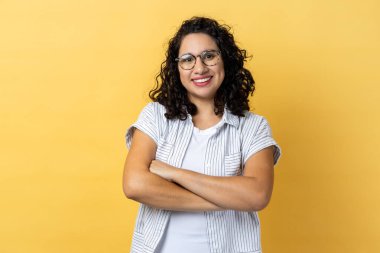 Çekici, mutlu, optik gözlüklü, siyah dalgalı saçlı, elleri katlı kameraya gülümseyen bir kadının portresi. Sarı arka planda kapalı stüdyo çekimleri.