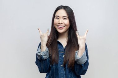 Gülümseyen, mavi kot ceketli, rock 'n roll jestli, neşeli, heavy metal dinleyen, gülümseyen bir kadının portresi. Gri arka planda izole edilmiş kapalı stüdyo görüntüleri.