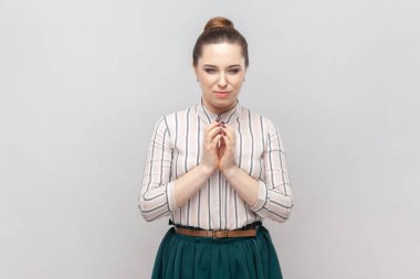 Çizgili tişört ve yeşil etek giyen kurnaz genç kadın portresi el ele tutuşuyor, iftira planları yapıyor, kafa yoruyor. Gri arka planda izole edilmiş kapalı stüdyo görüntüleri.