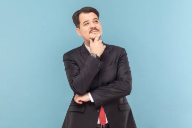 Kafası karışmış, kafası karışmış, bıyıklı, çenesini tutmuş, işini düşünen kırmızı kravatlı, siyah takım elbiseli bir adamın portresi. Açık mavi arkaplanda izole edilmiş kapalı stüdyo çekimi.